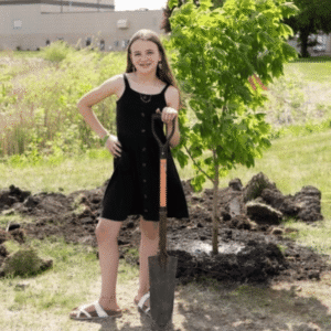 11-year-old Natalie Flaherty standing in front of planted tree sapling holding a shovel at the Fairmont Area High School