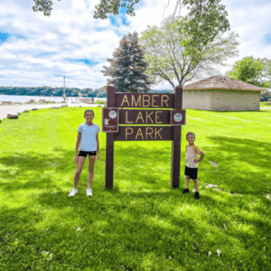 Two kids posing in front of the Amber Lake Park sign in Fairmont, MN.